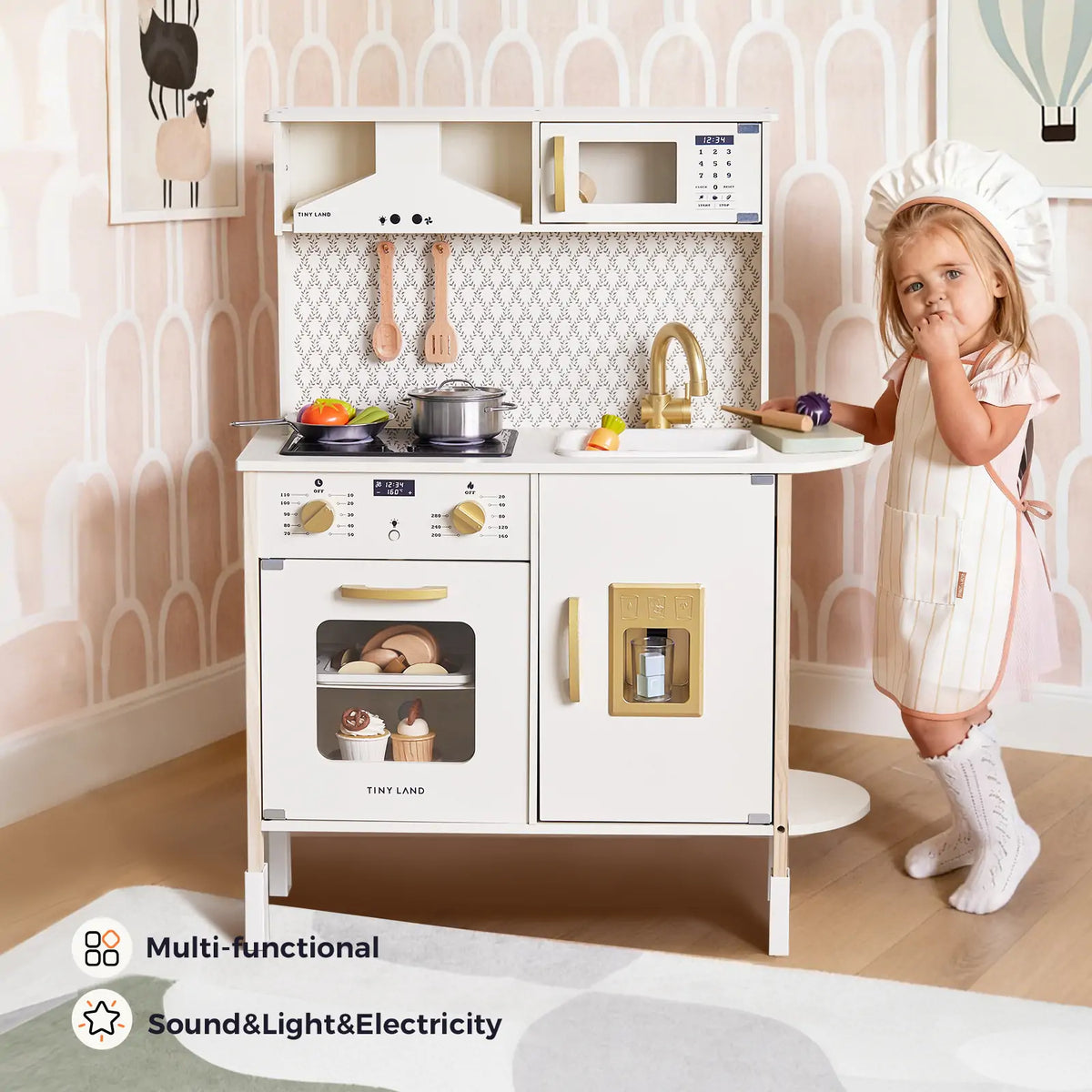 A young girl in a chef hat and apron stands by the Tiny Land® PopJoy Play Kitchen, which features gold accents, pots, utensils, appliances, and interactive lights and sounds—all in a playroom with patterned wallpaper.