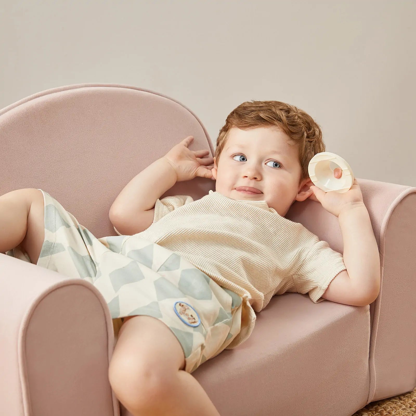 A young child with light brown hair relaxes on the Tiny Land® GentleSnug Kid Chair - Pink by Tiny Land, wearing a beige shirt and patterned shorts while holding a small round object and looking upward playfully.