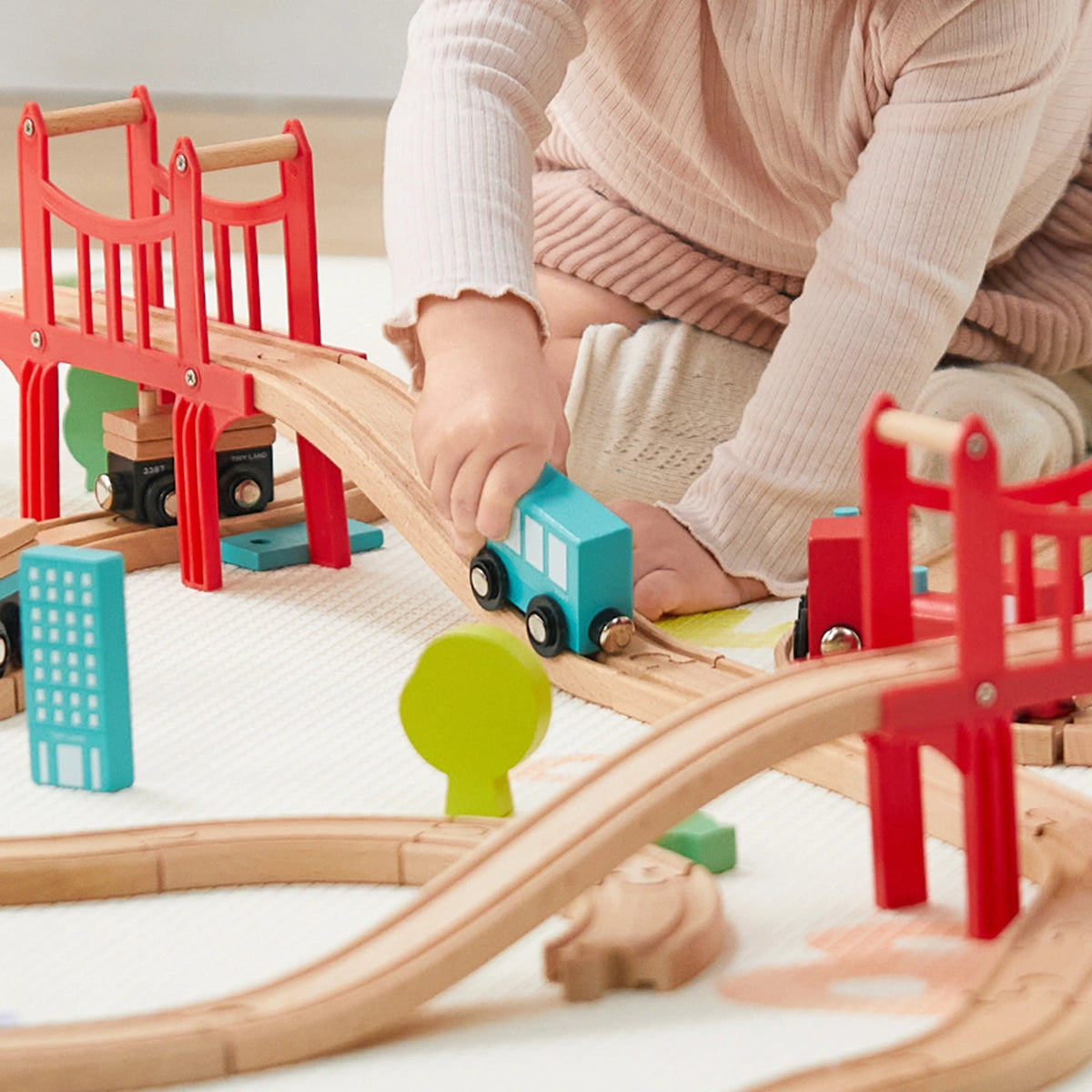 A child in a pink sweater and tights plays with the Tiny Land® Wooden Train Set for Children 39 Pcs, guiding a blue train over tracks and bridges, surrounded by toy buildings and trees.