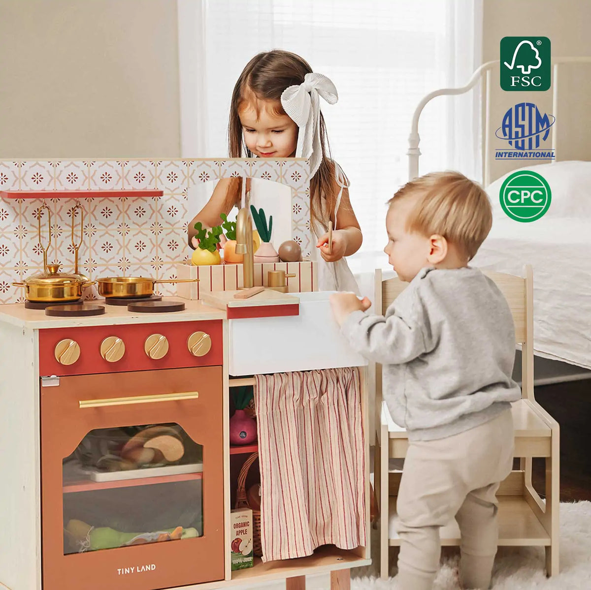Two young children play with the Tiny Land® Mediterranean Play Kitchen, pretending to cook with included pots, utensils, and play food. Tiny Land certification logos are visible in the upper right corner. A bed appears in the background|Mediterranean