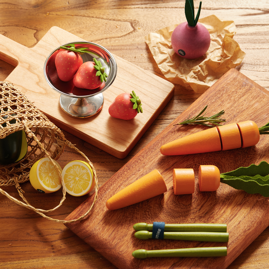 The Tiny Land® Organic Fruit & Veggie Pack by Tiny Land features realistic wooden strawberries, carrots, lemon, and a radish, artfully arranged on wooden boards and a table alongside toy tongs and a mesh bag—making it perfect as sensory produce toys or as part of a delightful wooden play food set.