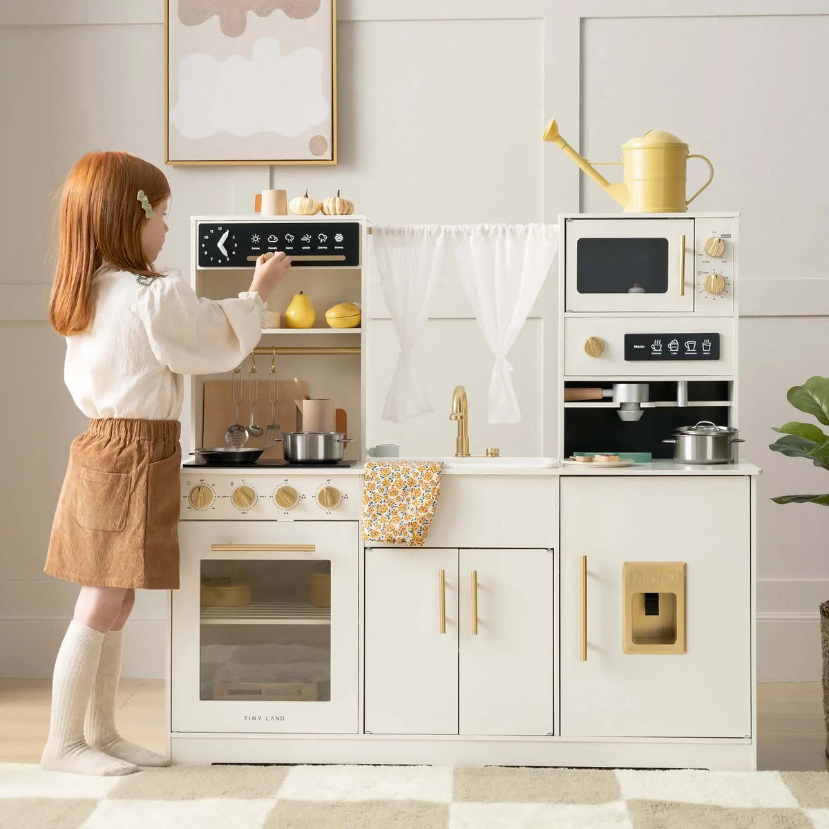 A young red-haired girl enjoys pretend play with the Tiny Land® Play Kitchen with Running Water - Iconic Aqua Kitchen in Cream, using the stove, oven, sink, and microwave from Tiny Land. Pots and a yellow watering can sit on top.