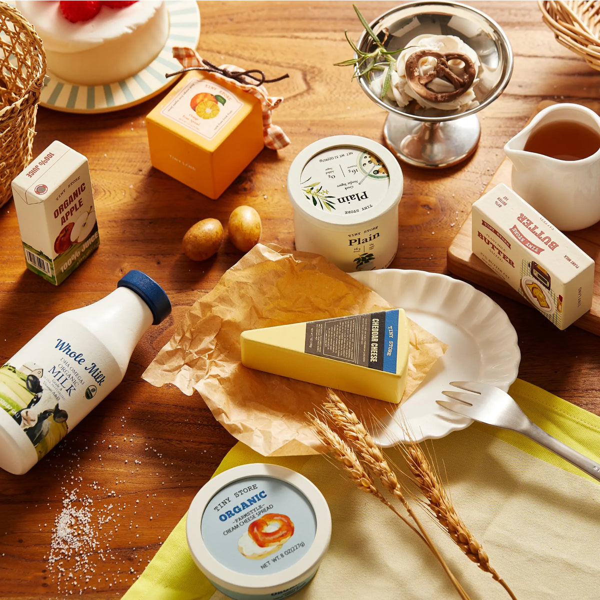 The Tiny Land® Organic Play Food - 4 Sets by Tiny Land features pretend cheese, milk, yogurt, cake, chocolate dessert, grains, and more on a wooden table—perfect for cozy, imaginative play with a rustic charm.