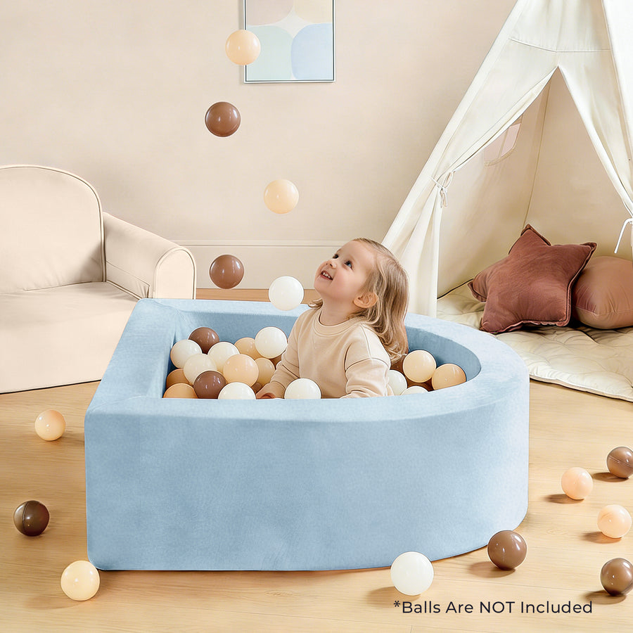 A young child grins while seated in a Tiny Land® PlayNest Soft Foam Ball Pit (Balls Not Included) in light blue, surrounded by brown and beige balls mid-air during sensory play. The background features a tent, sofa, climbing blocks, and pillows. Text reads, “*Balls Are NOT Included.”.|Blue