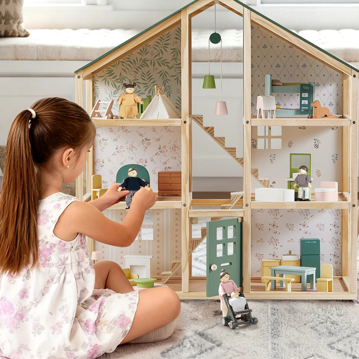 A young girl arranges Tiny Land® Wooden Dollhouse Dolls by Tiny Land INC in a cozy wooden dollhouse, placing them with miniature furniture on a light carpet in a bright, inviting room.