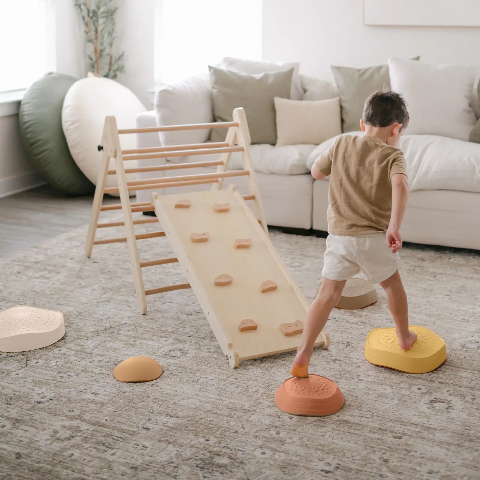 A young child balances barefoot on a yellow Tiny Land® Stepping Stone from the Sunset Hues set, navigating a living room obstacle course with more stepping stones, climbing equipment, cozy furniture, and natural light in the background.
