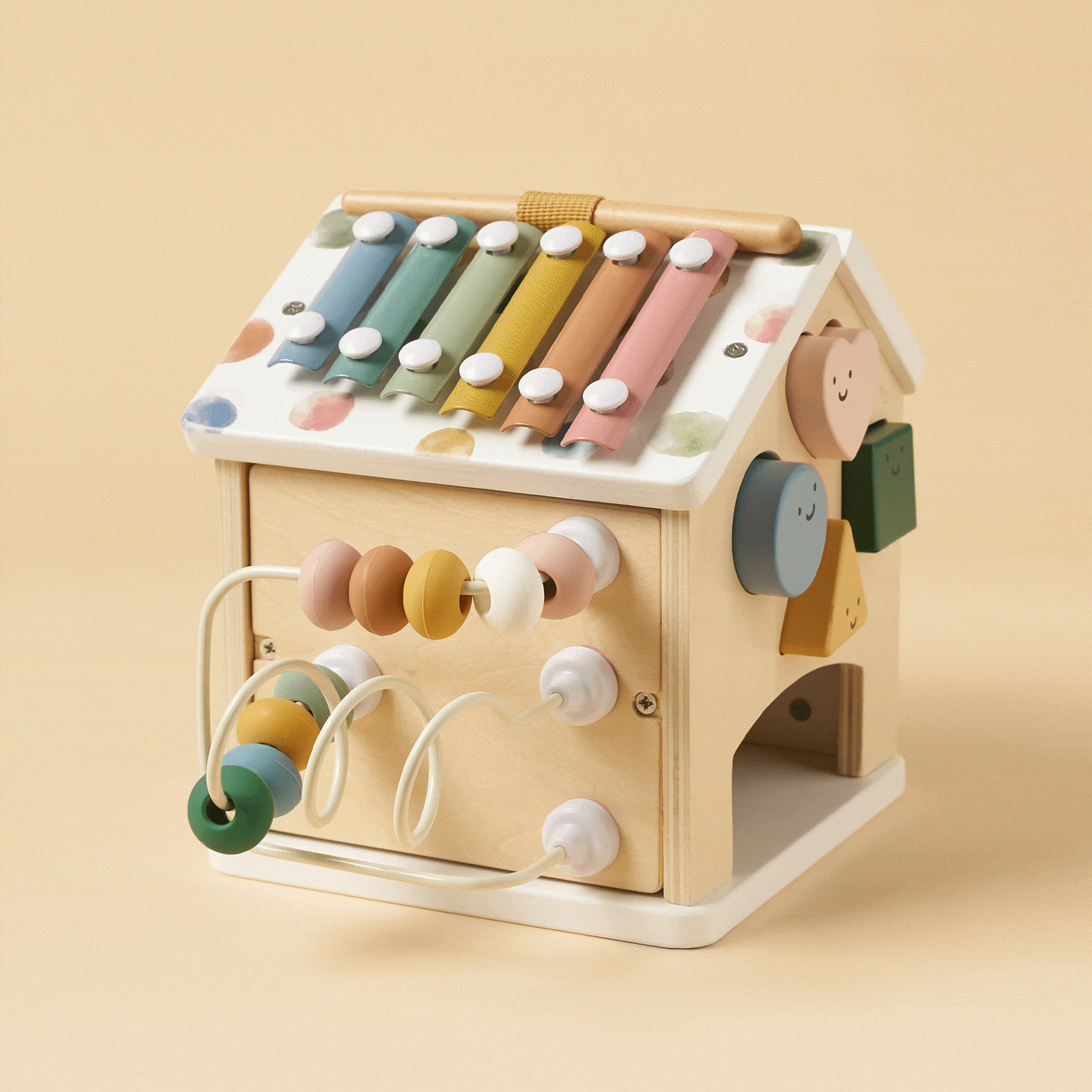 The Tiny Land® Cottage Activity Cube - Morandi by Tiny Land is a house-shaped wooden Montessori toy with a colorful xylophone, bead maze, spinning smiley gears, and pastel accents—an engaging developmental toy on a beige background.