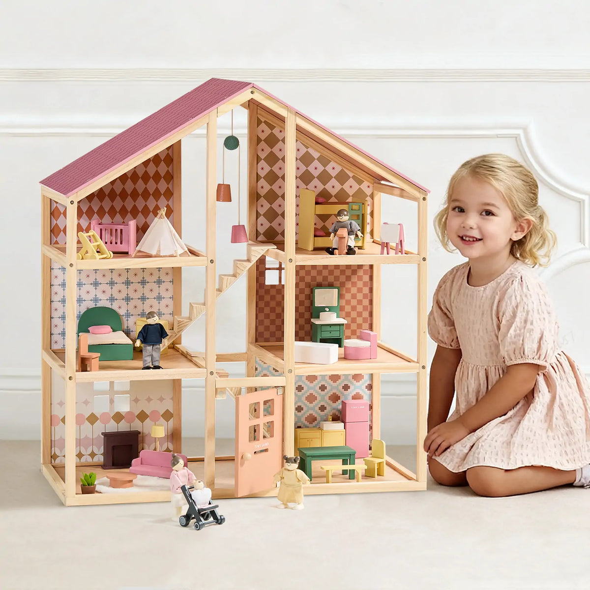 A young girl in a light pink dress sits beside the Tiny Land® Sweetwood Pegasus Dollhouse & Wooden Dollhouse Dolls by Tiny Land, featuring colorful mini furniture and poseable dolls, all set against a white wall with decorative molding.