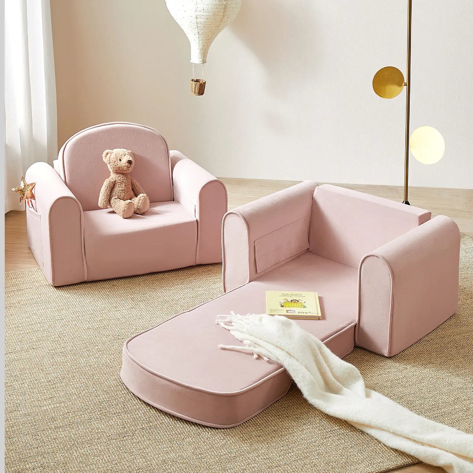 The Tiny Land® GentleSnug Kid Chair in pink sits beside a matching foldable kids’ sofa with a book and cream blanket, accented by a whimsical hot air balloon light—perfect playroom furniture for cozy, neutral rooms.