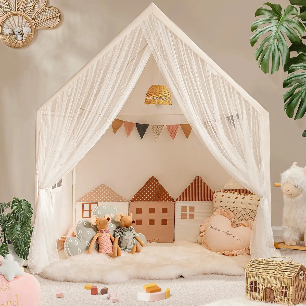 The Tiny Land® Chef’s Corner Playset features a cozy cream playhouse and kitchen set, complete with sheer white curtains, house-shaped pillows, stuffed animals, soft rugs, toys, and plants in a warmly decorated room.