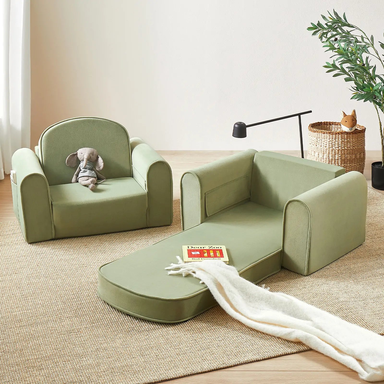 Two Tiny Land® GentleSnug Kid Chairs in sage sit on a beige rug; one upright holds a stuffed mouse, the other unfolds into a lounger with a book and white blanket. A plant and basket finish this cozy Tiny Land toddler seating area.