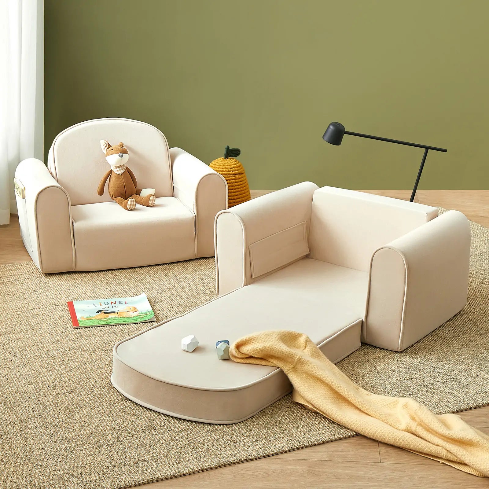 A cozy kids’ room features the Tiny Land® GentleSnug Kid Chair - Beige White, a stuffed animal, a foldable kids sofa bed with a yellow blanket, wooden blocks, a book on the rug, and a green basket against calming green walls.