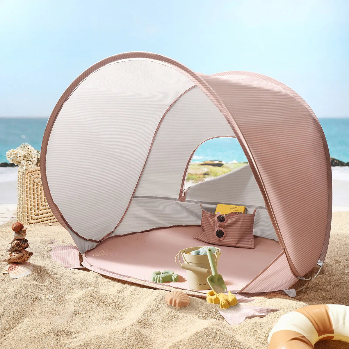The Tiny Land® One-Stop Beach Toy & Tent Set – Blush Pink features a small UV-protective tent and sand play set, perfect for the beach. It sits on sandy ground by the ocean, with a basket and decorative shells nearby under the blue sky.|Blush Pink