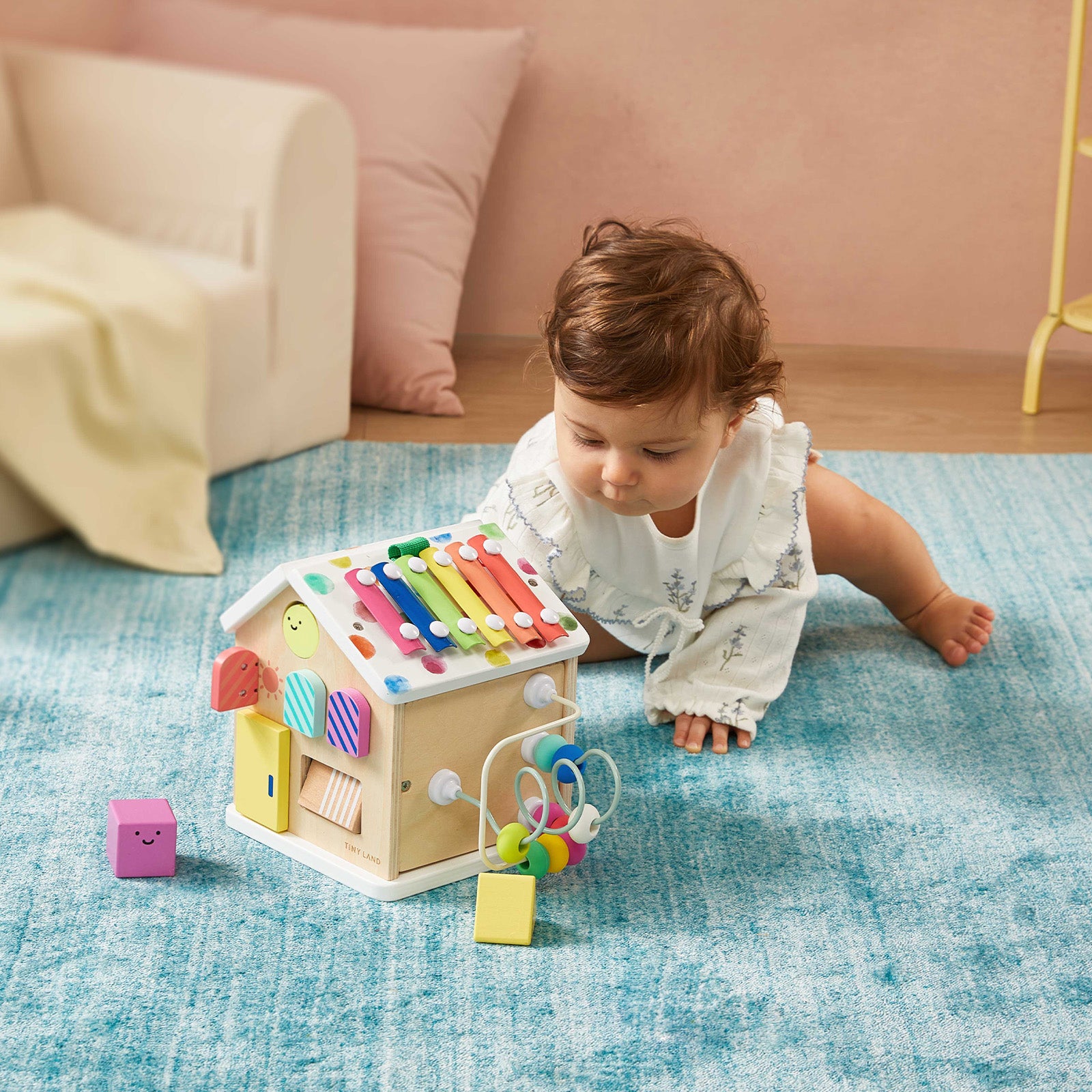 A baby in light clothes crawls on a blue rug, reaching for the Tiny Land® Cottage Activity Cube - Rainbow by Tiny Land, a house-shaped Montessori toy with musical bars, locks, and keys. Other small toys are scattered nearby.