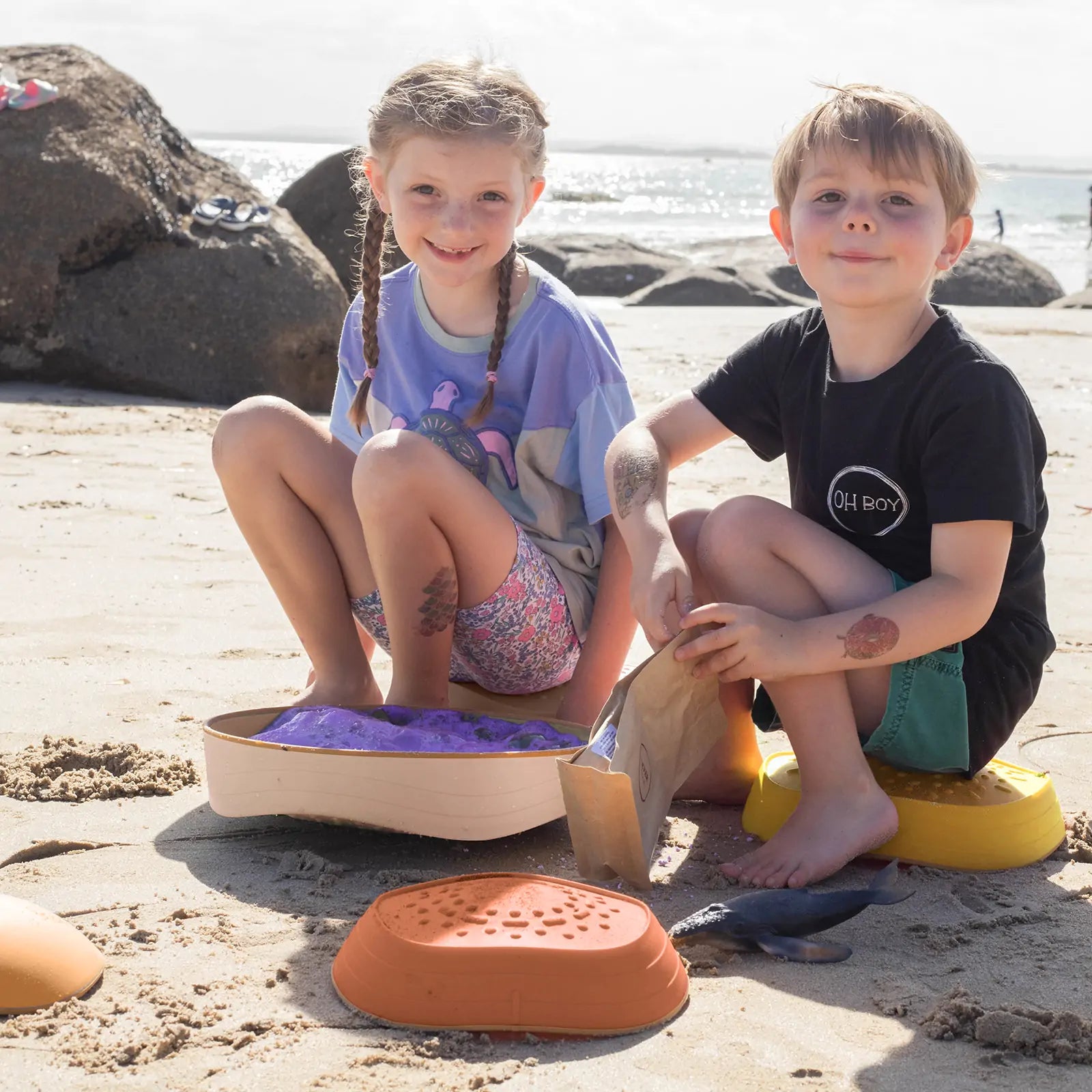Two young children play on the beach, smiling as they use Tiny Land® Stepping Stones - Sunset Hues to practice balance and coordination in the sunny weather, with rocks and the ocean in the background.