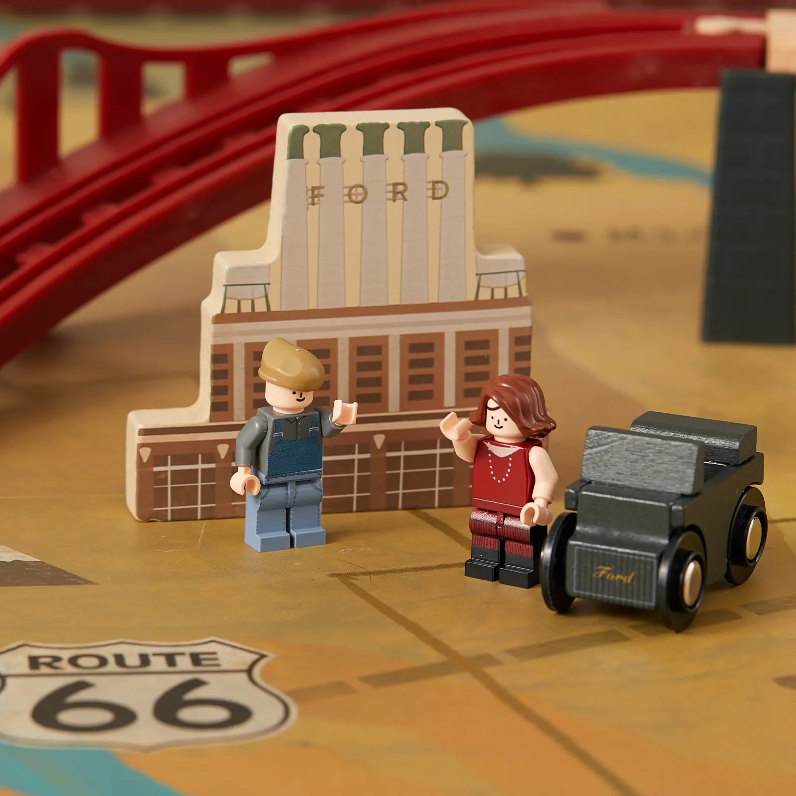 The Tiny Land® 1920 Railway Adventure Train Table by Tiny Land INC features toy figures, a wooden Ford building, a toy Ford car, an interactive city playset with a red bridge, map background, and Route 66 sign for imaginative adventures.