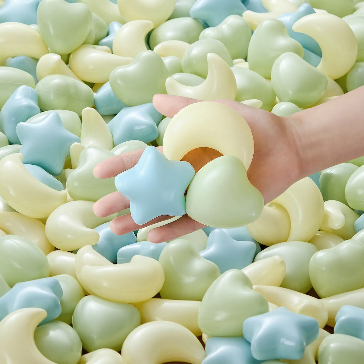 A hand holds pastel shapes—blue star, green heart, yellow crescent moon—above a pile of soft stars, hearts, and moons from the Tiny Land® Green Adventure 3-in-1 Play Tunnel & Fun-Shapes Ocean Ball Set by Tiny Land. Perfect for sensory play or ball pits.