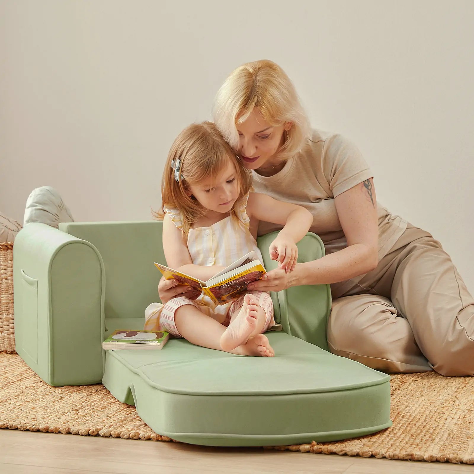A woman and a young girl read together on the Tiny Land® GentleSnug Kid Chair - Sage by Tiny Land. The barefoot girl sits on the extended seat while the woman points at a book, both enjoying a cozy, light-filled room.