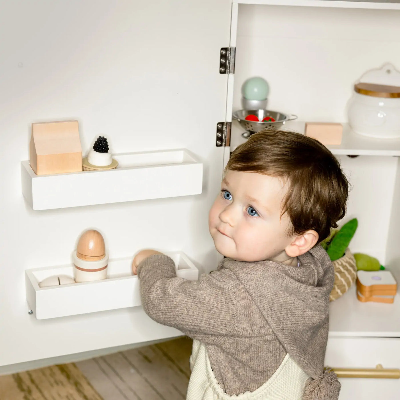 A young child with short brown hair and blue eyes reaches into the open Tiny Land® Play Kitchen with Fridge - Serenity Kitchen & Fridge Set in Cream, filled with toy food and wooden blocks for imaginative role-play fun.