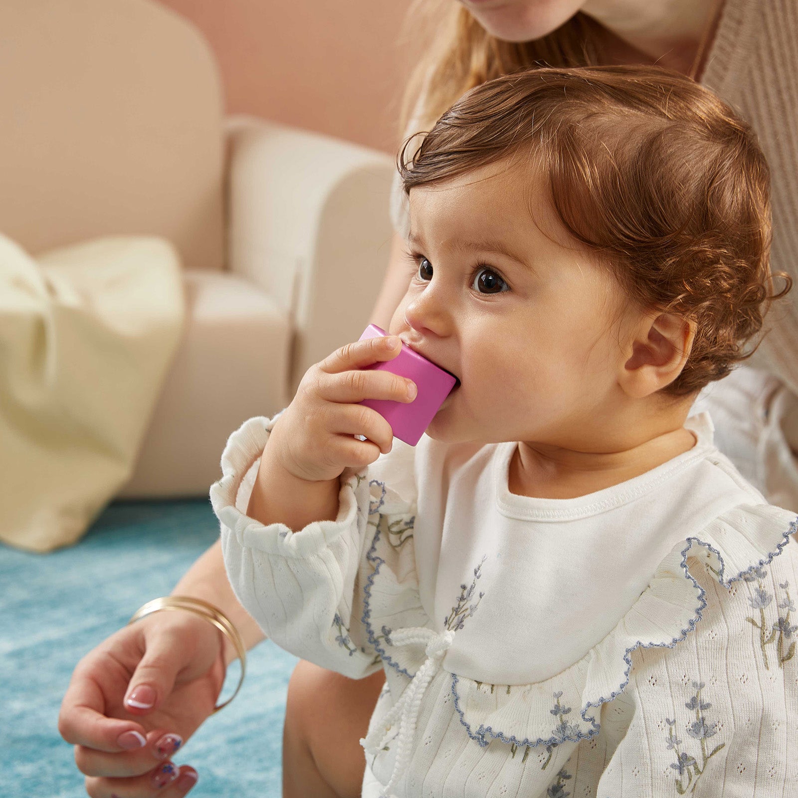 A toddler with curly hair in a white outfit sits on a blue carpet, holding the Tiny Land® Cottage Activity Cube - Rainbow by Tiny Land to their mouth, while an adult watches nearby.