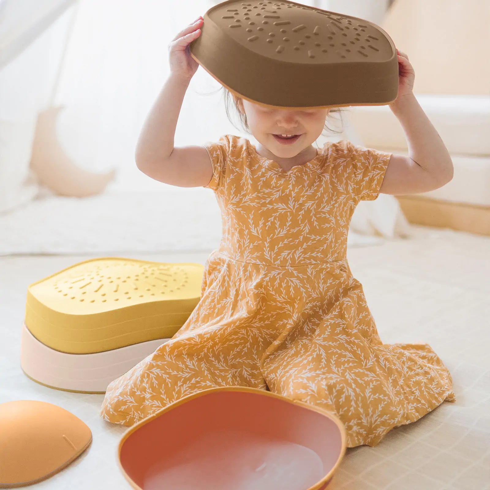 A young girl in a yellow floral dress smiles as she sits among colorful bowls, lids, and Tiny Land® Stepping Stones - Sunset Hues by Tiny Land, playfully holding a brown bowl over her head for coordination and balance fun.