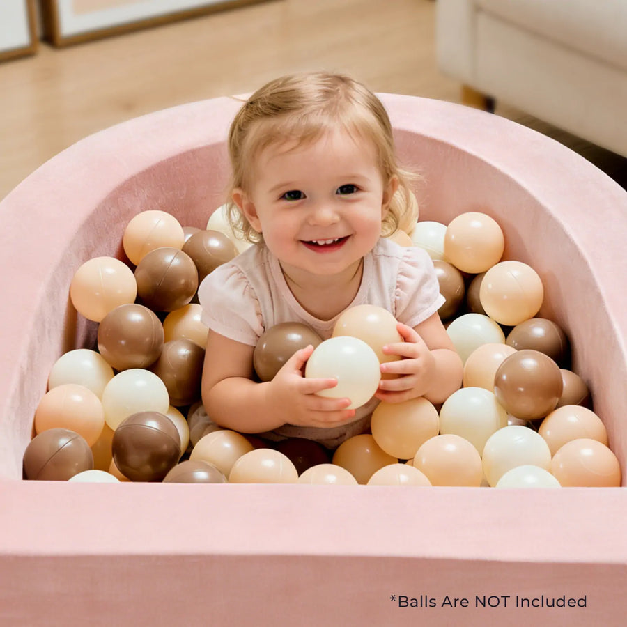 A smiling young child sits in the Tiny Land® PlayNest Soft Foam Ball Pit by Tiny Land, surrounded by cream, beige, and brown balls (balls not included), enjoying engaging sensory play. A note at the bottom reads, “*Balls Are NOT Included.”.|Pink