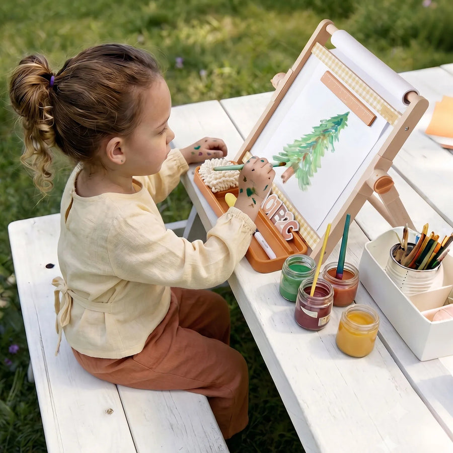 A young child sits at a white picnic table outdoors, painting a tree on the Tiny LandĀ® Double-Sided Magnetic Tabletop Easel. Various paint jars and brushes from Tiny Land are arranged beside her on the table, creating a vibrant art station.|Oatmeal