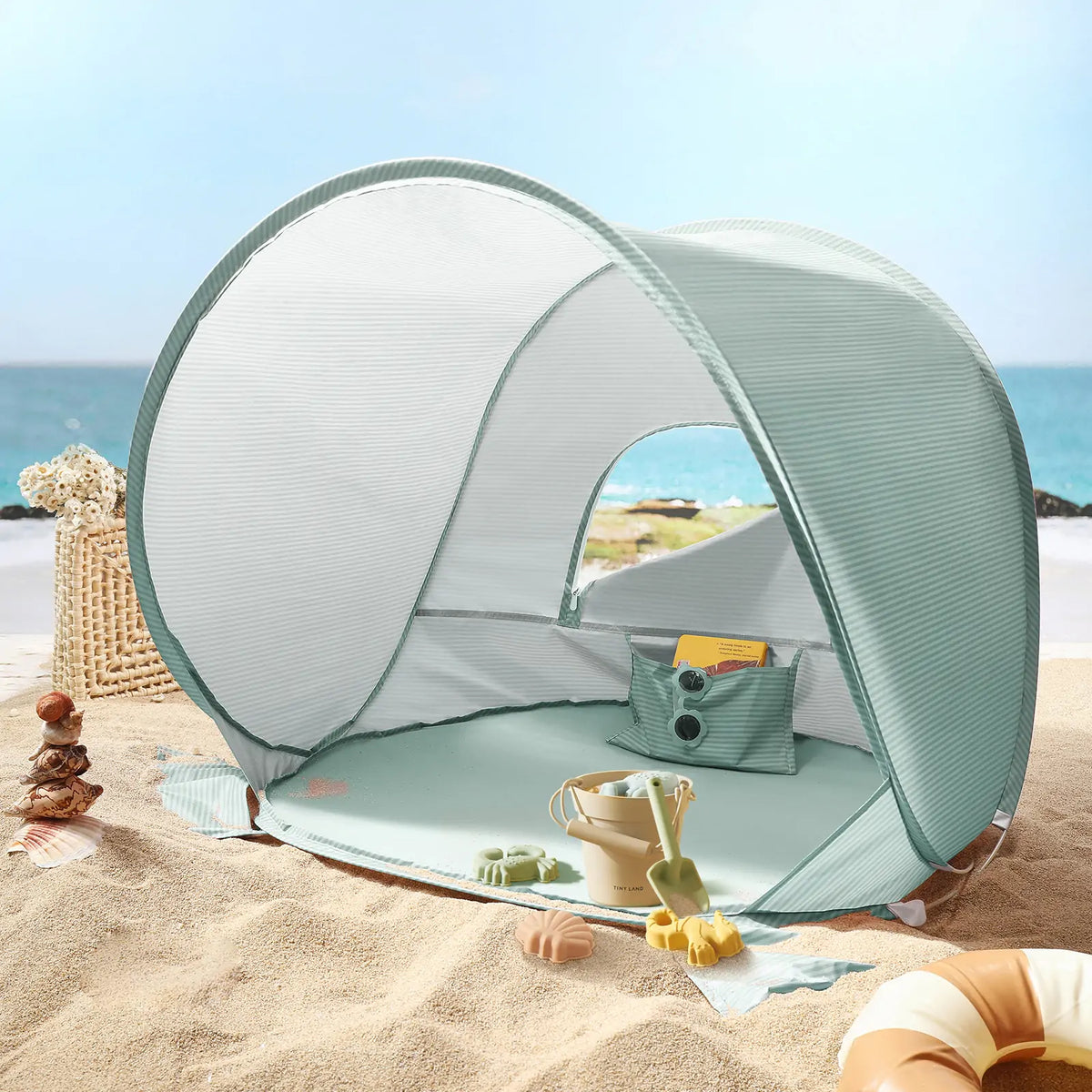 The Tiny Land® One-Stop Beach Toy & Tent Set – Sea Blue provides UV-protective shade on the sand, with included digging tools and a wicker basket nearby, all set against a serene sea and blue sky.|Sea Blue