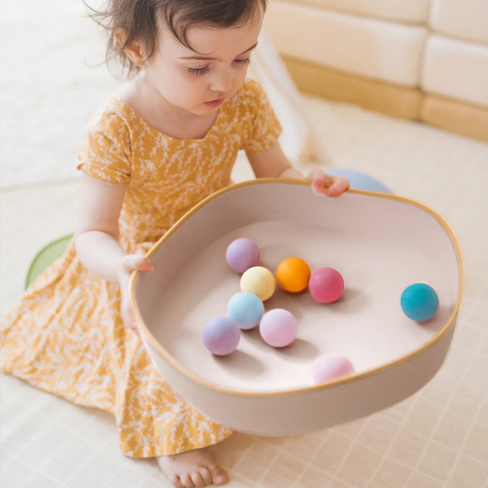 A young child in a yellow dress sits on the floor with a bowl of colorful balls. Nearby, Tiny Land® Stepping Stones - Sunset Hues by Tiny Land add fun to the play area while encouraging balance and coordination.