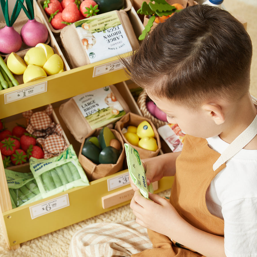 A child in a tan apron holds a green packet while standing by a play grocery display filled with the Tiny Land® Organic Play Food Collection from Tiny Land Play Food—realistic toy fruits, vegetables, and cartons that perfectly mimic a market setting.