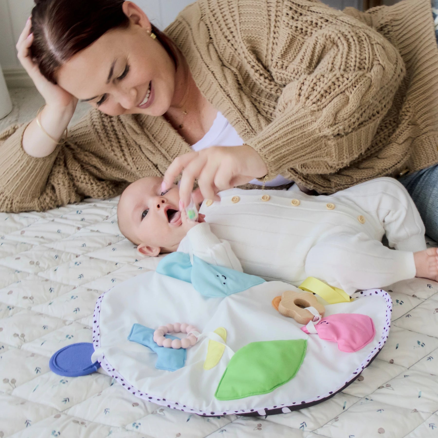 A woman in a brown sweater smiles while playing with her baby, dressed in a white onesie and enjoying Tiny Land® Montessori Toys for Newborns (0–3 Months) from the Tiny Land Official Store on a colorful playmat.