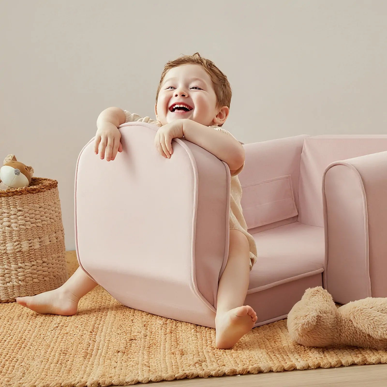 A smiling child sits playfully on the Tiny Land® GentleSnug Kid Chair in pink, with a beige woven rug, a stuffed bear, and a basket of toys in the background.