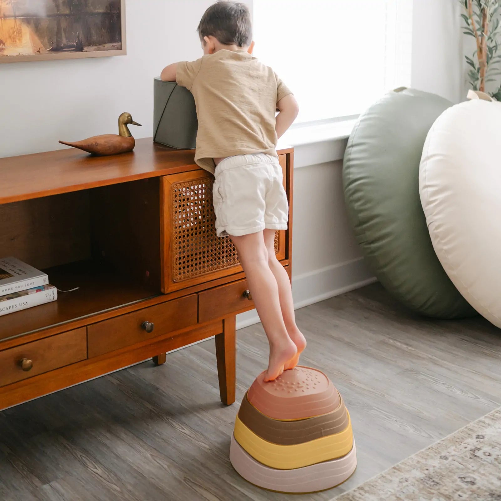 A young child uses Tiny Land® Stepping Stones - Sunset Hues by Tiny Land to build balance and coordination while reaching for a box atop a wooden cabinet in a bright, modern living room.