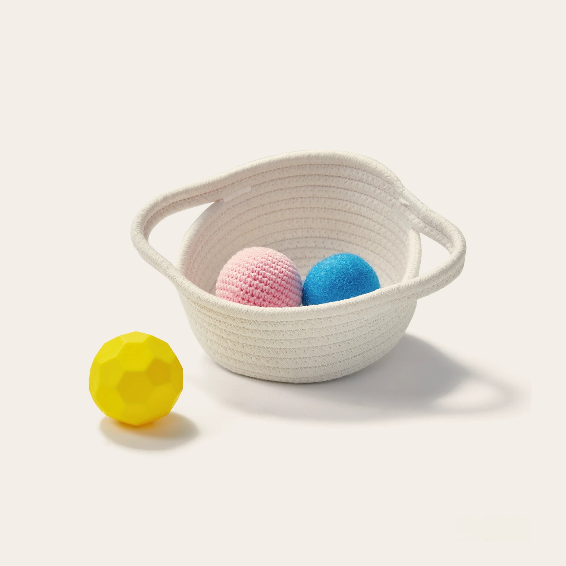 A white woven basket holds a pink knitted ball and a blue felt sensory ball, with a faceted yellow ball beside it—all from Tiny Land® Montessori Toys for Newborns (7–9 Months) by Tiny Land, perfect for early sensory play.