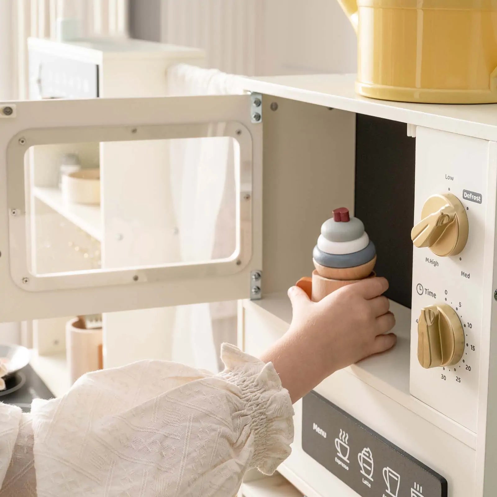 A child in a white long-sleeve shirt puts a toy ice cream cone into the Tiny Land® Play Kitchen with Running Water—Iconic Aqua Kitchen in Cream, featuring control knobs and a yellow teapot, bringing pretend play to life.