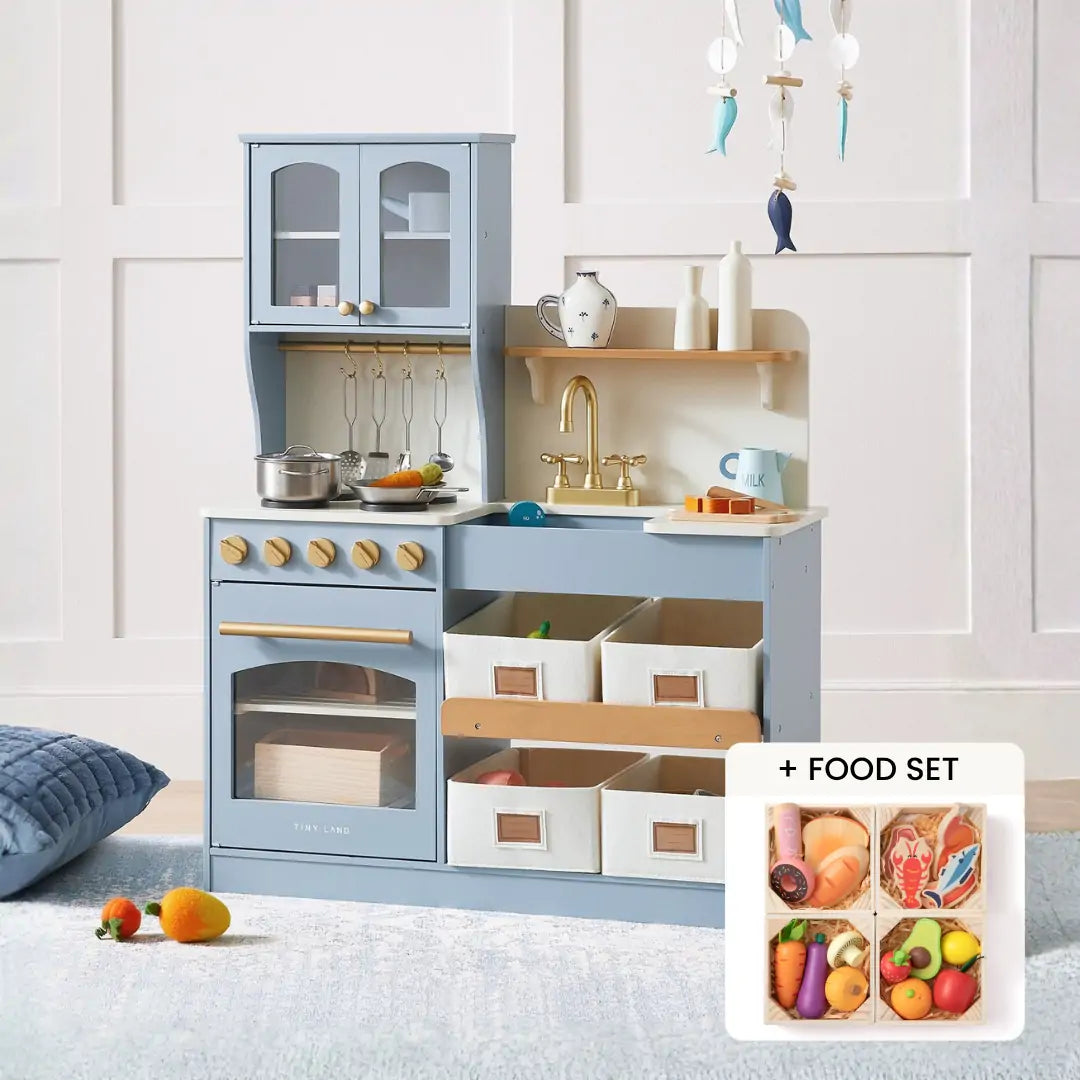The Tiny Land® Serenity Wooden Kitchen & Wooden Food Set by Tiny Land features a light blue kitchen with shelves, bins, sink, and stove. It comes with utensils, pots, and colorful pretend food. Shown against a white wall with a nearby cushion.