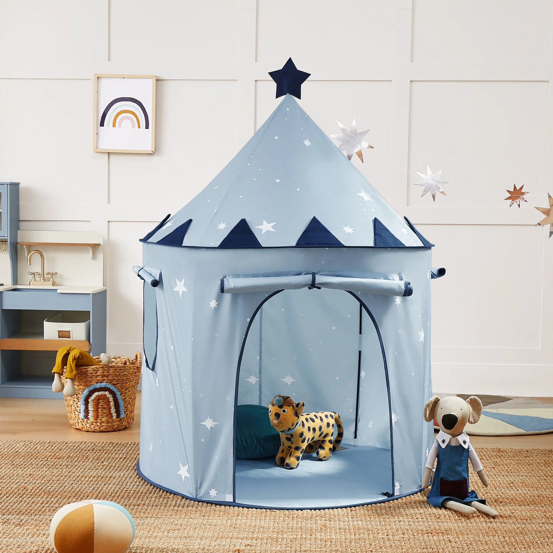 The Tiny Land® Kids Tent – Starry Blue Indoor Tent for Toddlers stands in a children’s room. Inside are a stuffed leopard and blue pillow, while a plush dog, toys, and rainbow decor rest on a tan rug nearby.