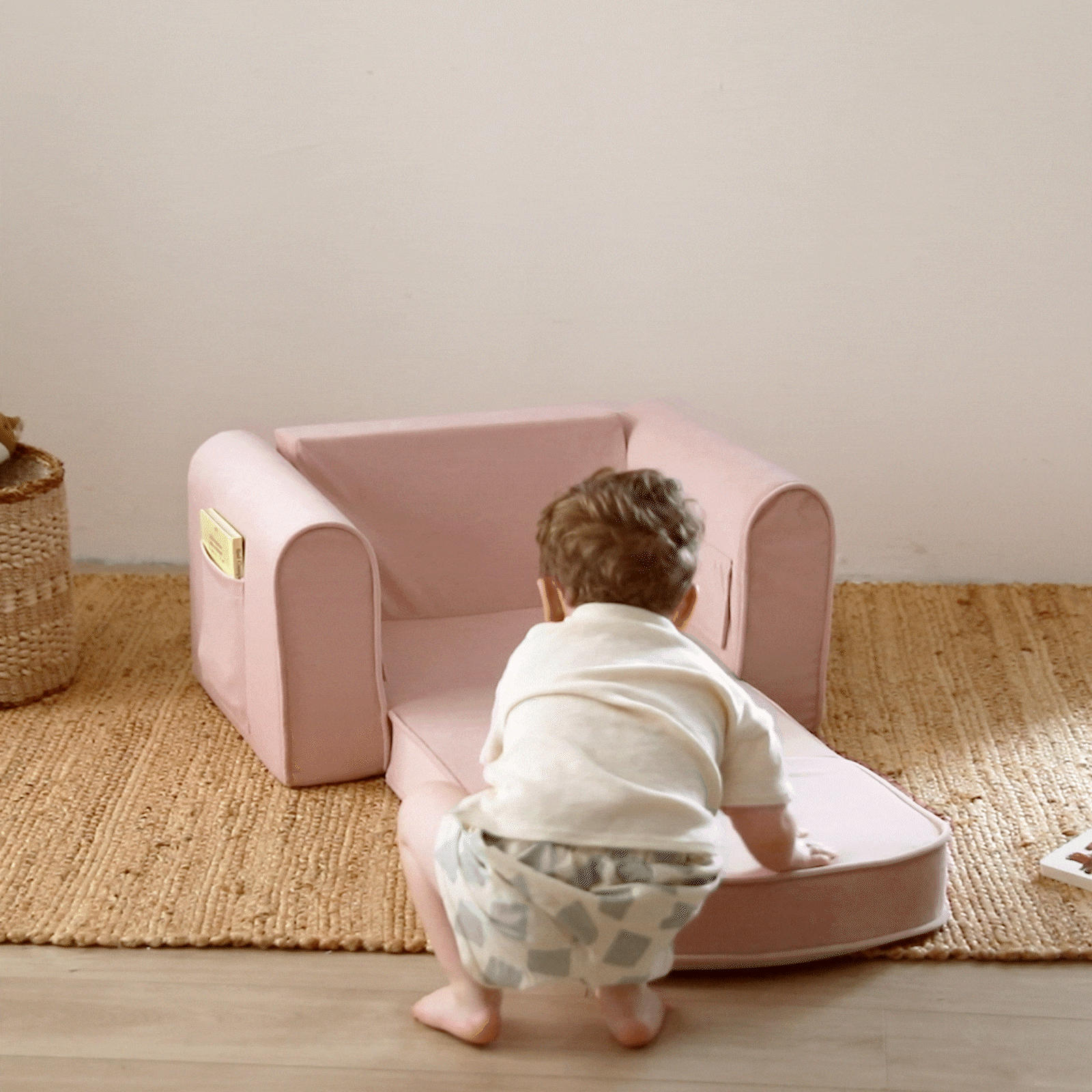 A toddler in a white shirt and patterned shorts kneels on a woven rug, playing with the Tiny Land® GentleSnug Kid Chair - Pink by Tiny Land, which features a soft foldable design with a leg rest—ideal playroom furniture.