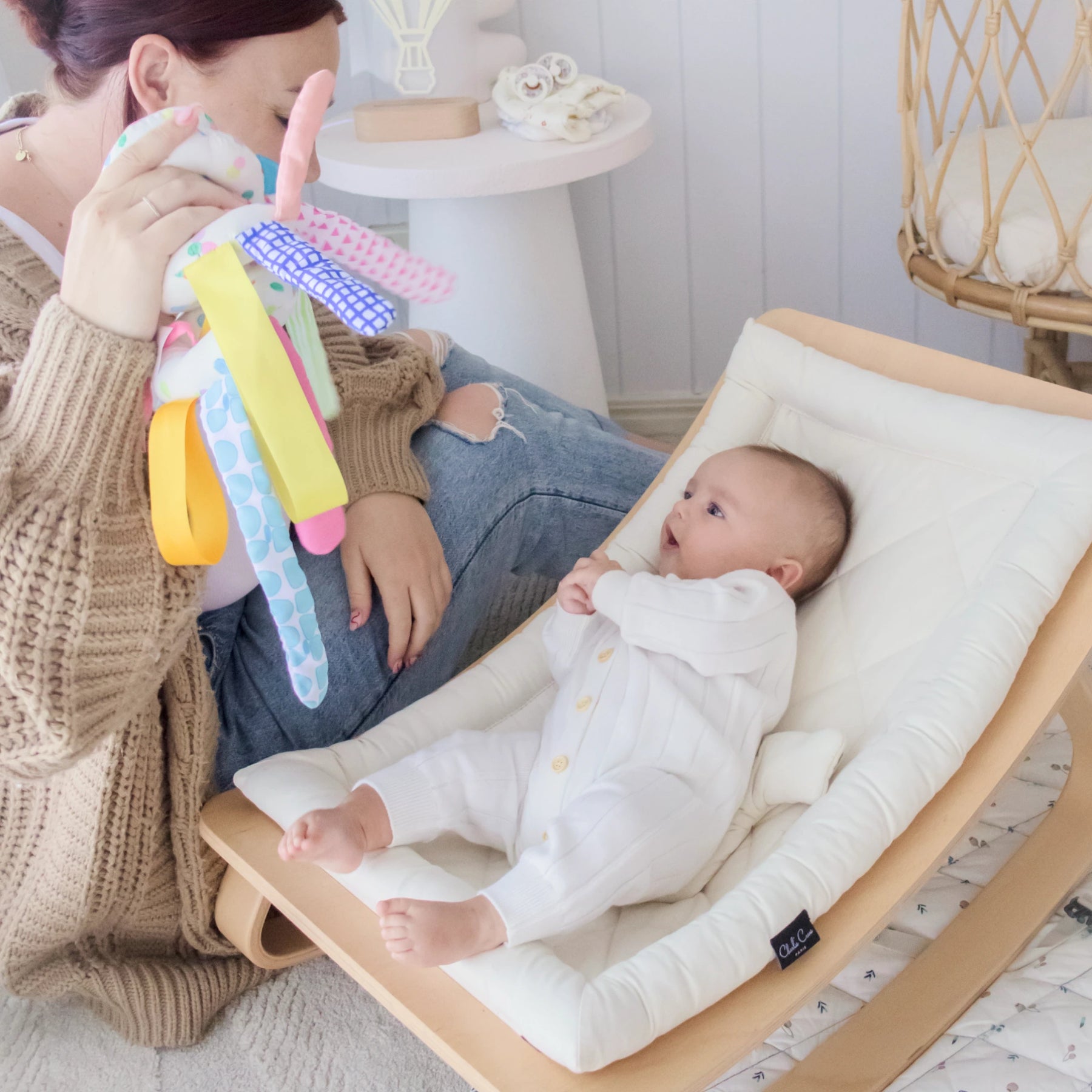 A woman holds a Tiny Land® Montessori Toy for Newborns from the Tiny Land Official Store above a baby in a cream lounger. The baby, in a white onesie, looks up with curiosity, enjoying early sensory play designed for ages 0–3 months.