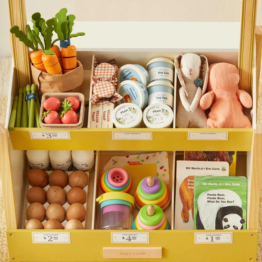 The Tiny Land® Kid's Grocery Store Playset from Tiny Land features a yellow market stand with plush veggies, wooden eggs, toy jars, stacking cups, a stuffed bunny, and children’s books—all neatly arranged for imaginative play.