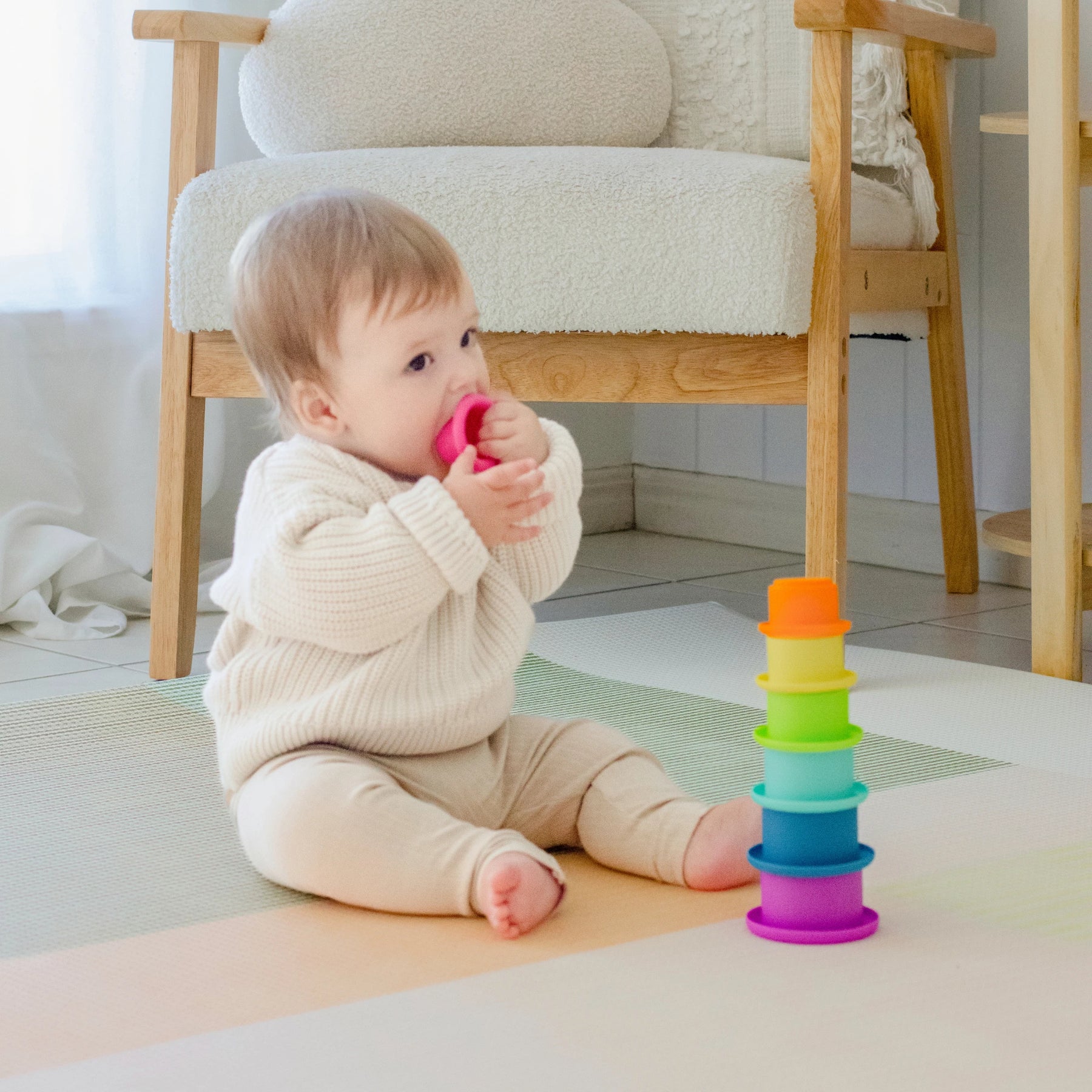 A baby in cream clothes sits on the floor, holding a pink stacking cup from the Tiny Land® Montessori Toys for Newborns (7–9 Months) by Tiny Land. Colorful cups, a sensory ball, and a wooden chair are nearby.