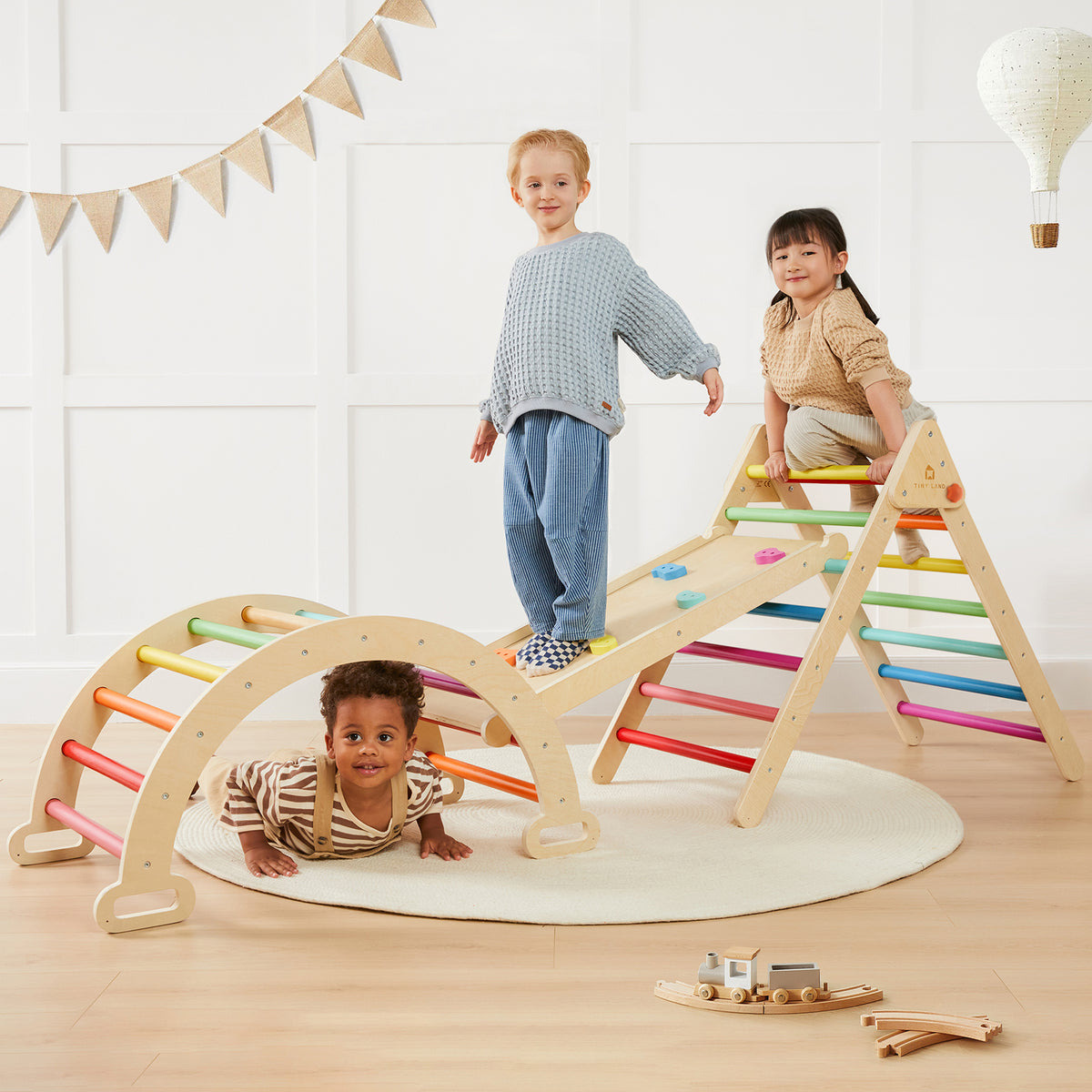 Three children enjoy the Tiny Land® 7-in-1 Rainbow Climbing Set in a bright room: one stands on the balance board, another climbs the Pikler Triangle, and a third crawls under an arch. A toy train and balloon decor create a playful atmosphere.|rainbow color