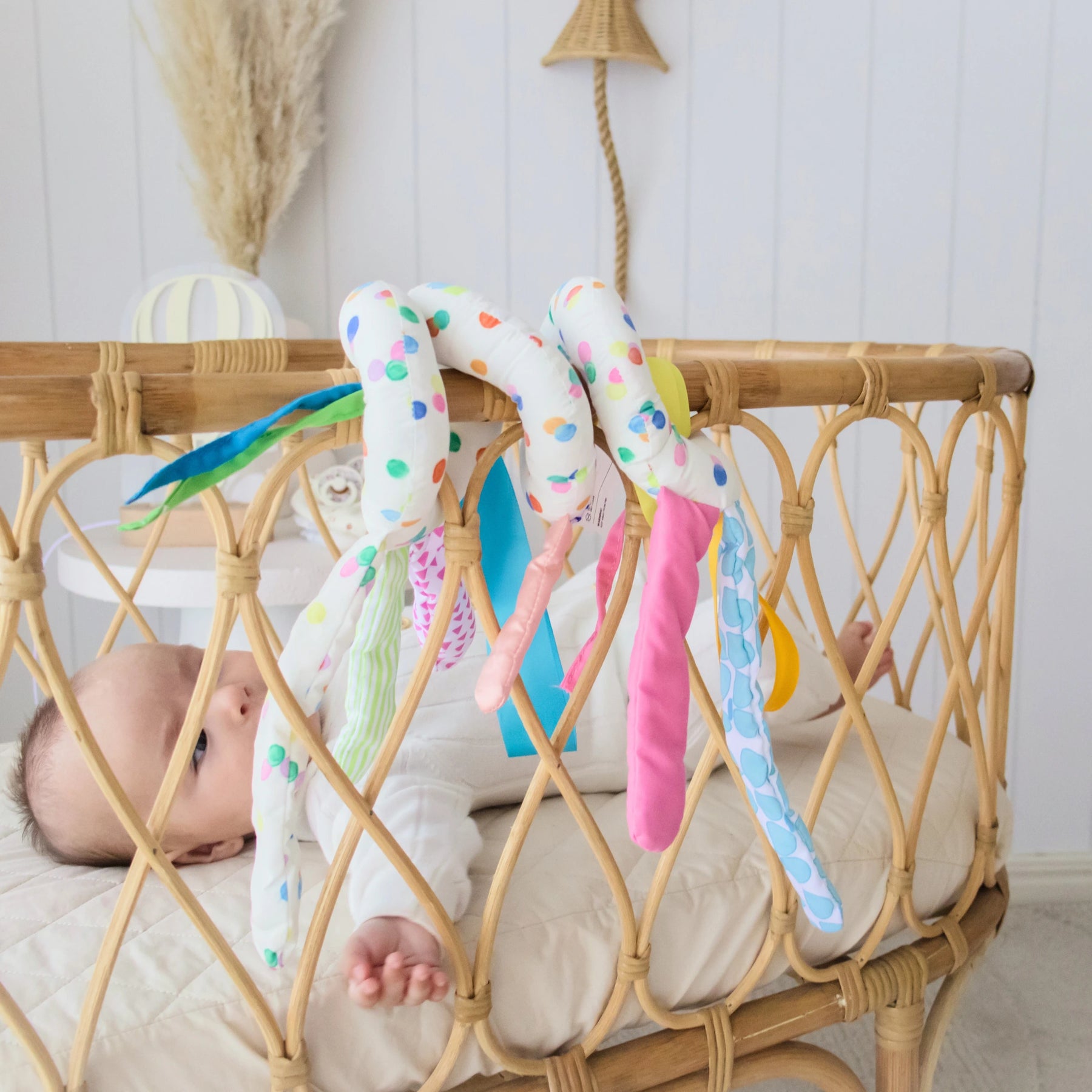 A baby lies in a wicker crib, reaching for soft, colorful fabric snakes from the Tiny Land® Montessori Toys for Newborns (0–3 Months) by Tiny Land Official Store. The nursery has light walls and natural decor.