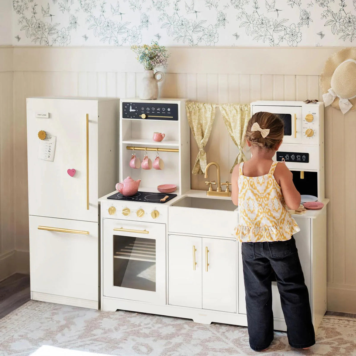 A child in a yellow and white top plays with the Tiny Land® Premium Play Kitchen & Fridge Set by Tiny Land—which features a kids’ play fridge, stove, sink, and microwave—in a bright, decorated room.
