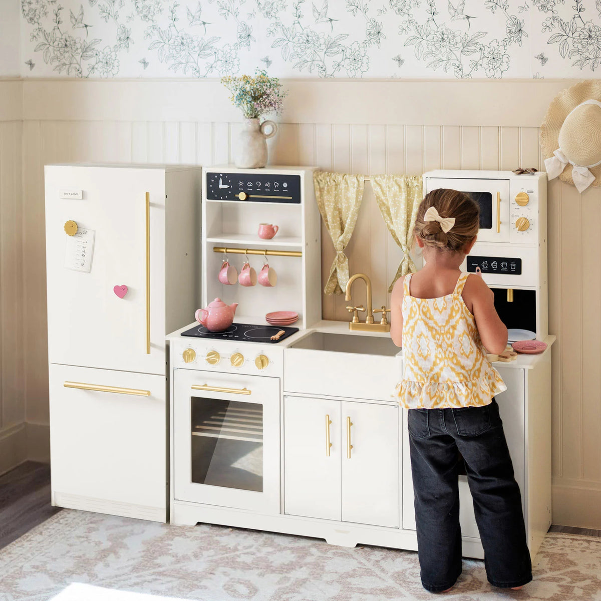 A young child enjoys pretend play with the Tiny Land All-in-One Family Feast Gift Set: Luxury Kitchen & Fridge & Organic Food Collection, surrounded by floral wallpaper and light furnishings for an inviting play space.