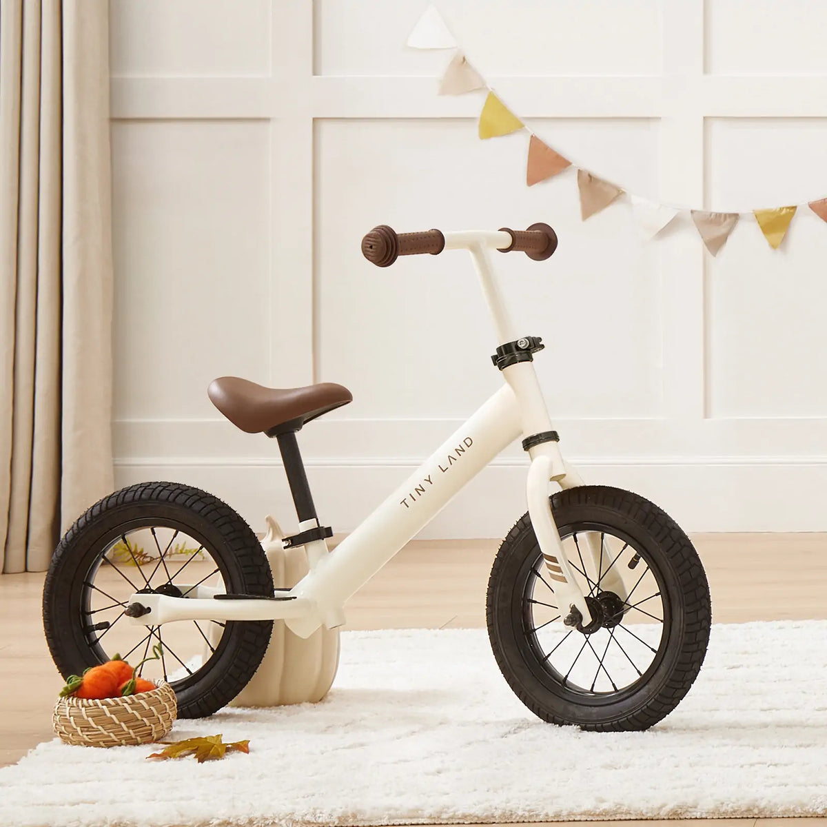 The Tiny Land® Balance Bike - Milky White, featuring black rubber tires and a brown adjustable seat, is shown indoors on a wooden floor. A woven basket with small pumpkins sits nearby under decorative bunting on a white wall.