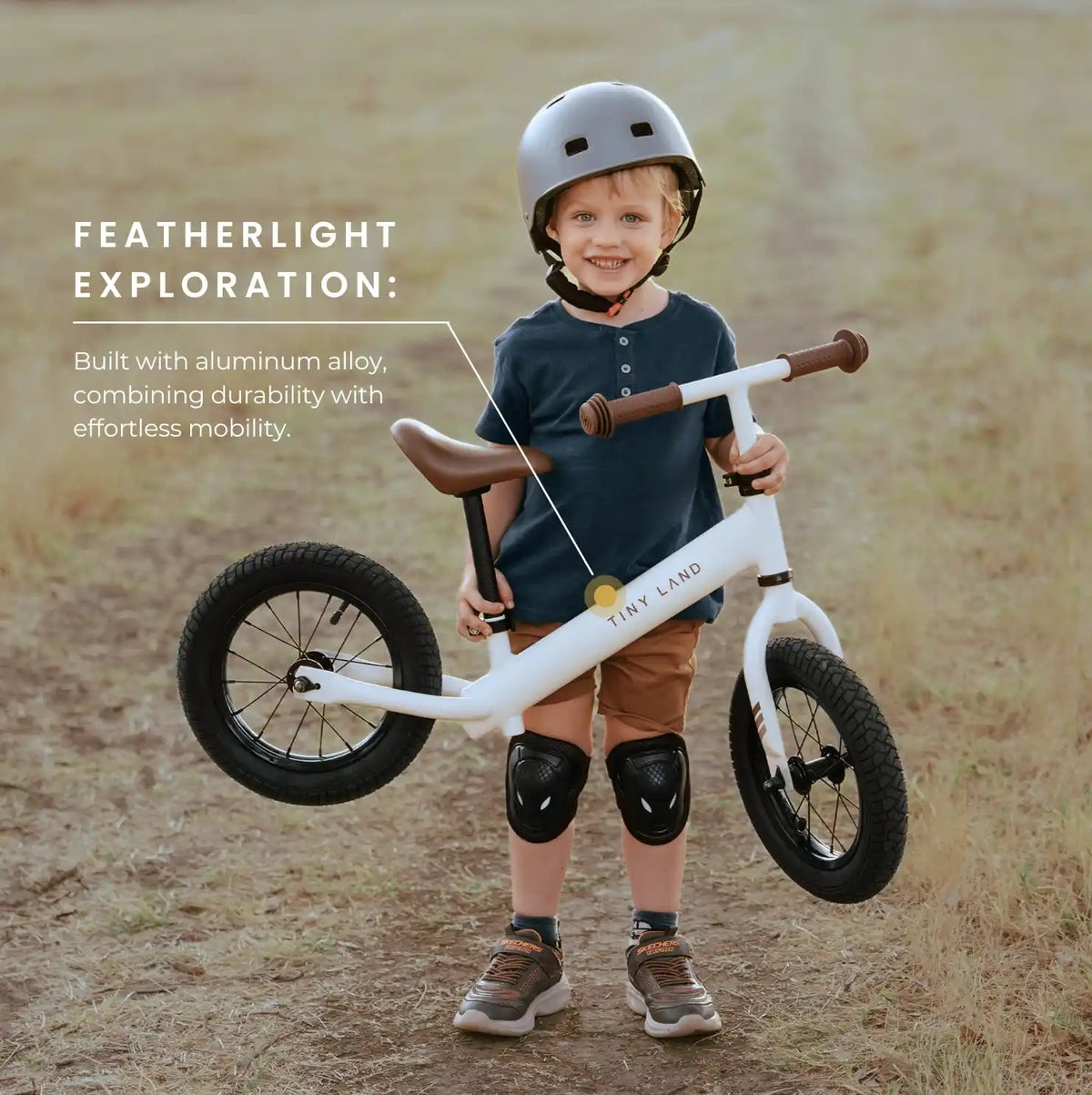 A smiling child in a helmet and knee pads stands on a dirt path with the Tiny Land® Balance Bike - Milky White, featuring a lightweight aluminum alloy frame, adjustable seat, and durable rubber tires for comfort and easy mobility.