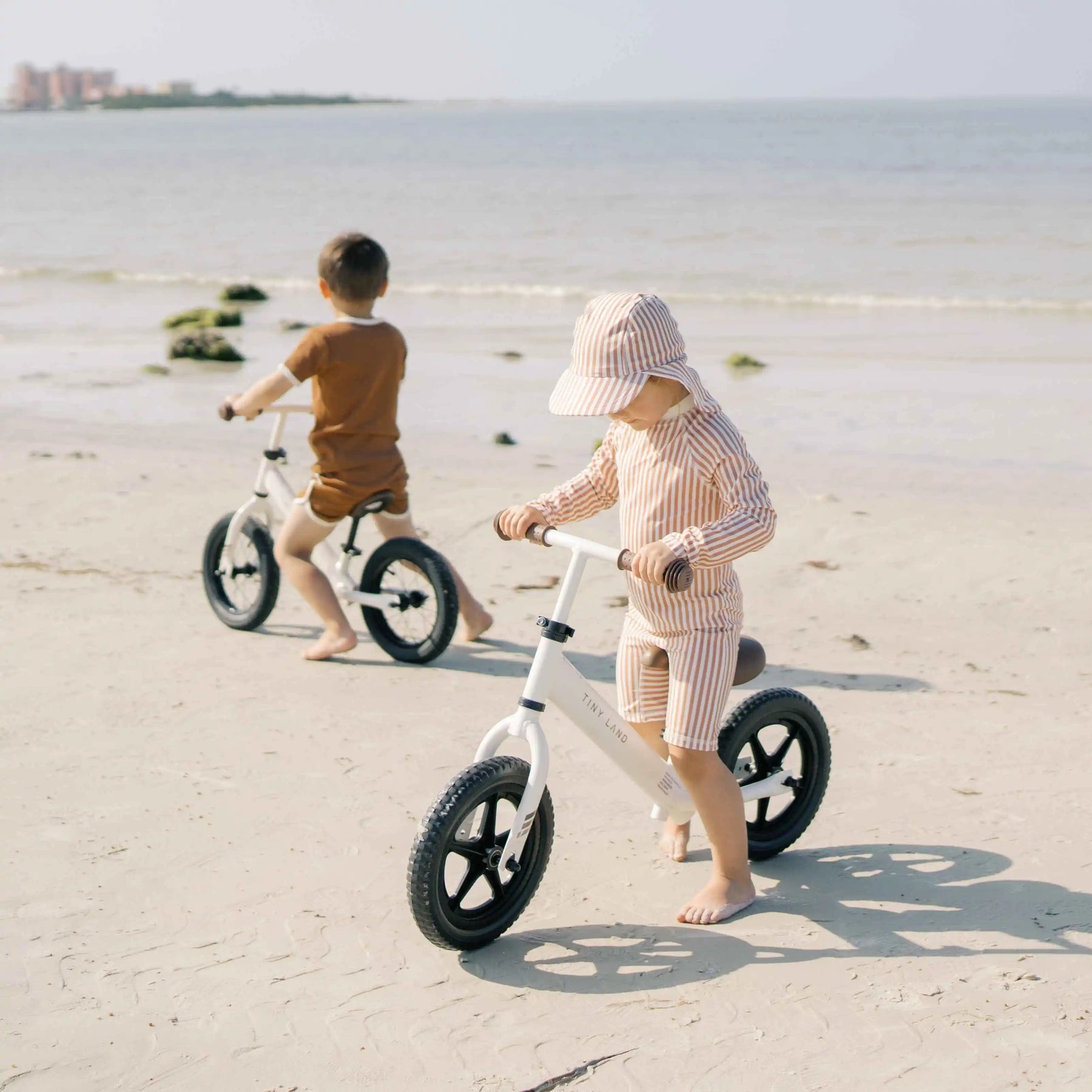 Two young children ride Tiny Land® Balance Bike - Milky White with adjustable seats and rubber tires barefoot on a sandy beach near the water. One wears stripes and a hat; the other, brown shorts and a t-shirt. The ocean frames the scene.