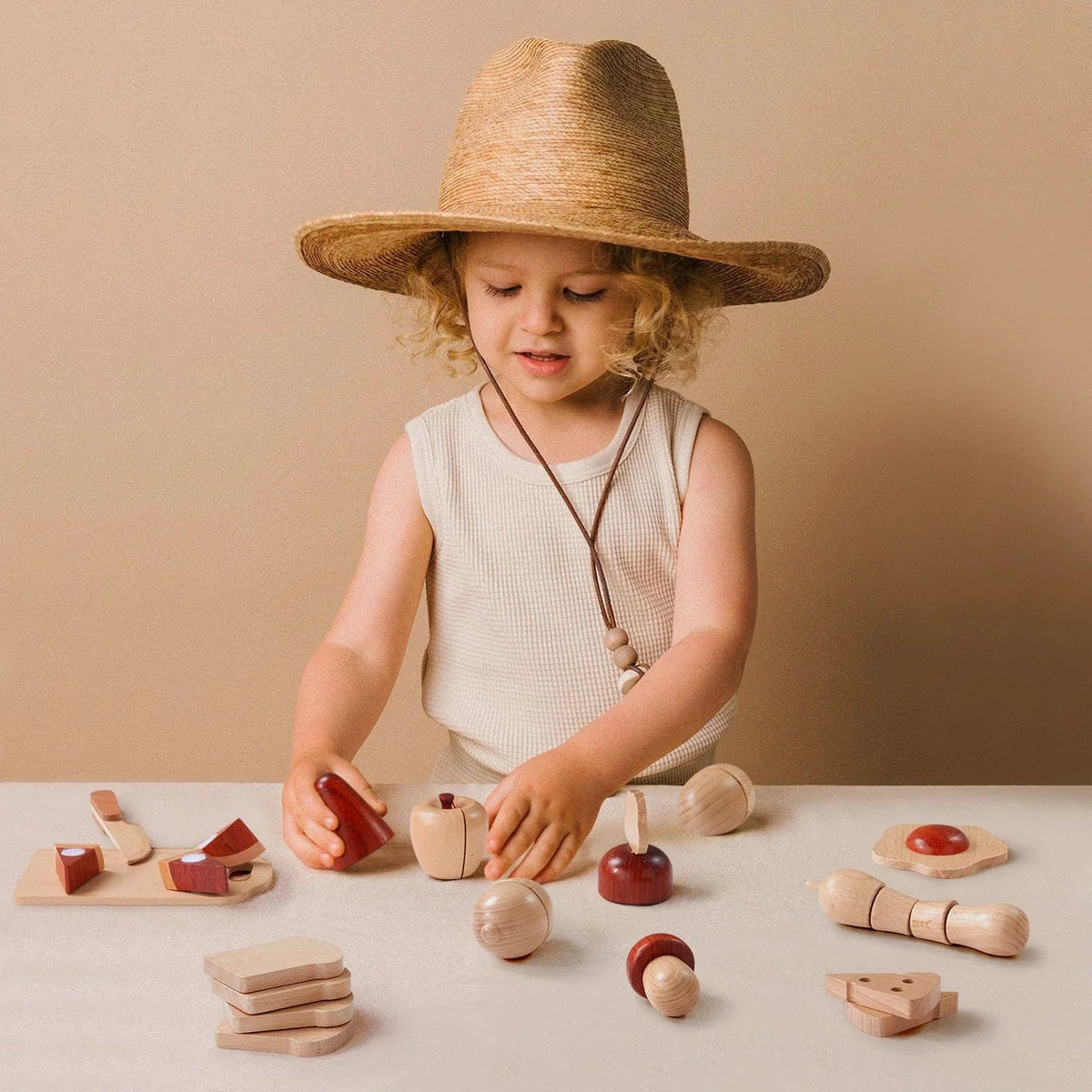 A young child with curly blond hair in a straw hat happily plays with Tiny Land® Play Kitchen Accessories – Wooden Cut and Play Food Toys on a beige table against a matching background.