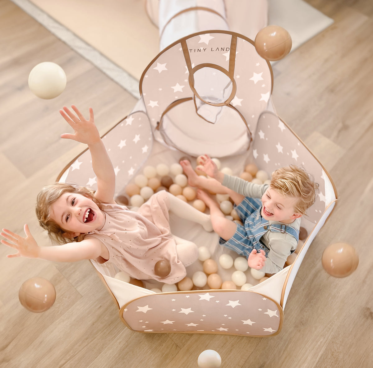 Two young children laugh and toss Tiny Land® Ocean Ball - 200 pcs with Varied Colors in a star-patterned, beige playpen filled with soft, non-toxic balls. A play tunnel attaches to the playpen on a light wood floor. Brand: Tiny Land.|Brown